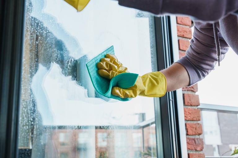 A person wearing yellow gloves wipes a glass window with a sponge, removing soap and streaks for a clear view outside