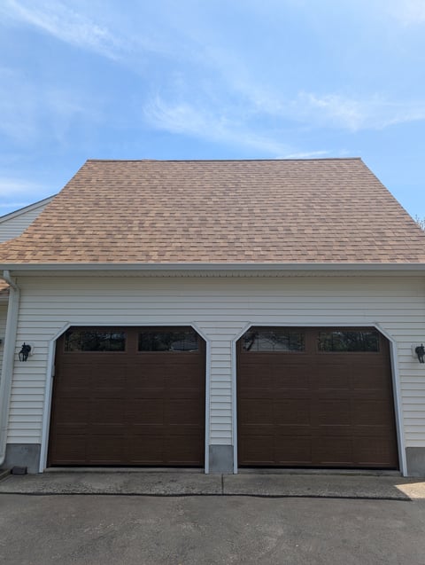 Two-car garage with brown doors, white siding, and shingled roof under blue sky
