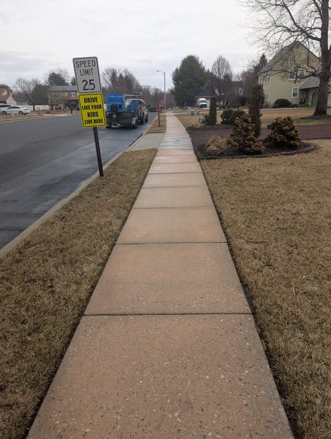 Straight concrete sidewalk alongside a residential street with a speed limit 25 sign and parked vehicles visible