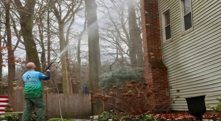 Person in blue shirt using garden hose to spray water in residential yard with bare trees and brick house