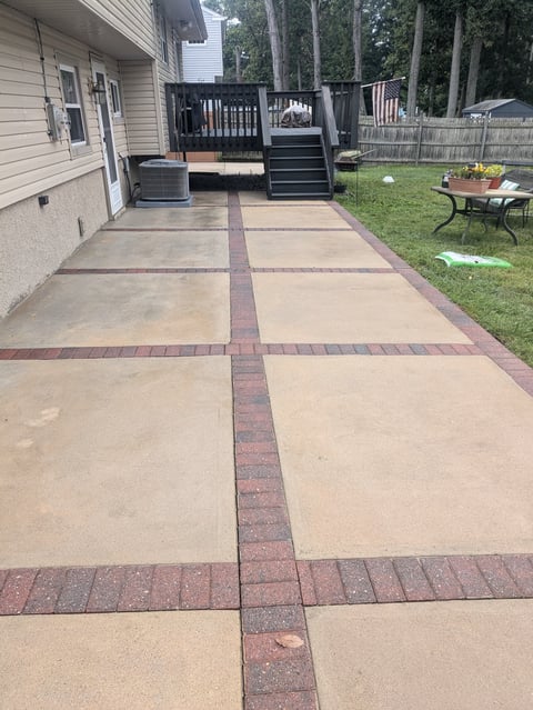 Residential backyard with paved patio featuring decorative brick border pattern, leading to a black deck and grassy area with trees
