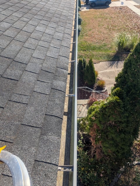 Aerial view of asphalt shingle roof with metal gutter, overlooking landscaped yard with evergreen shrubs and parked car