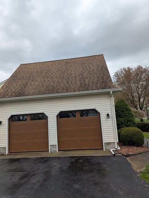 Two-car garage with brown doors on white siding, shingled roof, asphalt driveway, and landscaping under cloudy sky